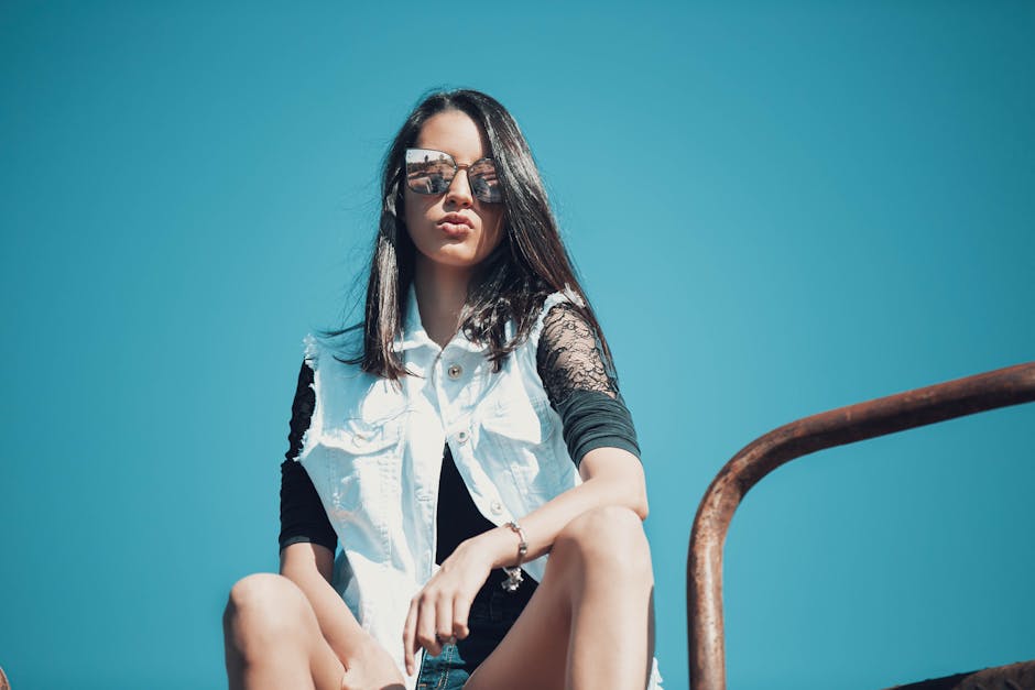 Fashionable young woman sitting outdoors in sunglasses and white vest posing confidently under clear blue sky.