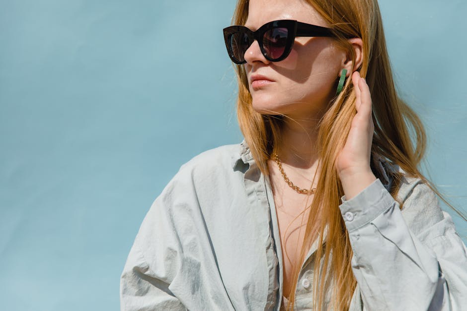 Stylish woman with blonde hair wearing sunglasses and a casual shirt against a blue background.