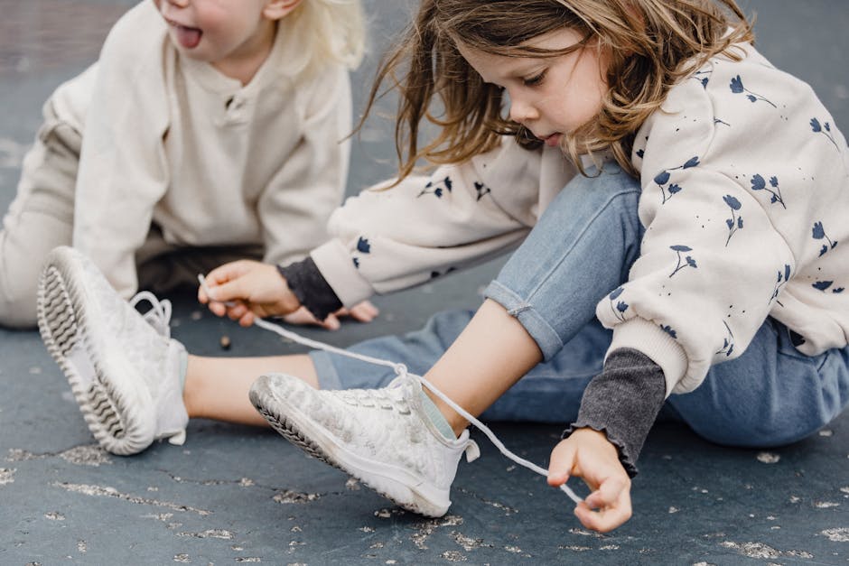 Concentrated little girl tying laces on sneakers while sitting with bent leg near anonymous kid with blond hair and tongue out