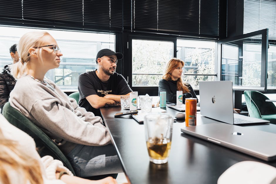 A group of adults engaged in a discussion during a team meeting in a modern office with laptops and refreshments.